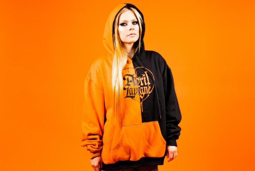 IG/avrillavigne/Avril Lavigne dengan hoodie oranye dan hitam bertuliskan namanya.